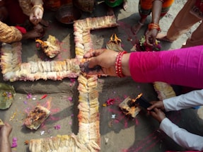 A group of people gather around a cross-shaped arrangement made of woven ropes or fibers, adorned with flowers and colored powders. Some are holding incense sticks and performing a ritual by lighting small fires on the arrangement.