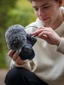 A person is adjusting a microphone attached to a camera. They are holding the camera with one hand while using the other hand to handle the microphone. The person is wearing a light-colored hoodie and appears focused on their task.