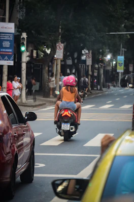 A motorcycle taxi driver helping a passenger with a helmet on a sunny city street.