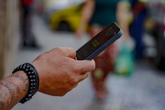 A tattooed hand wearing a black beaded bracelet is holding a smartphone. The background is blurred, showing people walking on what appears to be a city street.
