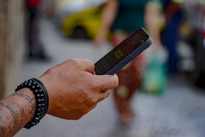 A tattooed hand wearing a black beaded bracelet is holding a smartphone. The background is blurred, showing people walking on what appears to be a city street.