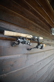 Two fishing rods with cork handles and spinning reels are mounted on a wooden wall. The wooden surface has a rich, dark brown tone with visible grain patterns and a slightly glossy finish.