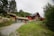 A rustic wooden cabin with a grass roof is situated on a hillside surrounded by lush greenery and trees. A larger red house with a dark roof and a chimney is also present. The area is landscaped with stones, creating a natural and quaint environment.