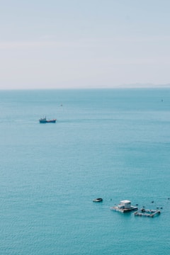 A tranquil, expansive view of the ocean with a distant ship on the horizon. Near the foreground, floating structures likely used for aquaculture are visible, surrounded by calm blue-green water.