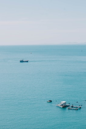 A tranquil, expansive view of the ocean with a distant ship on the horizon. Near the foreground, floating structures likely used for aquaculture are visible, surrounded by calm blue-green water.