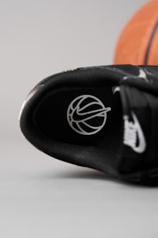 A close-up view of the inner part of a black sneaker, featuring a logo resembling a basketball with white lines. In the background, a portion of an orange basketball can be seen.