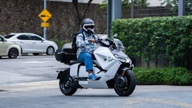 A person wearing a helmet and colorful jacket rides a sleek, modern white electric scooter on a city street. The background features greenery and a stone wall, with a couple of stationary cars in the vicinity, indicating an urban setting.