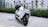 A sleek, modern electric scooter with a white and black color scheme, parked on a paved area next to green shrubs and red-tinted plants. The design is angular with a prominent front light and visible handlebars.