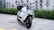 A sleek, modern electric scooter with a white and black color scheme, parked on a paved area next to green shrubs and red-tinted plants. The design is angular with a prominent front light and visible handlebars.
