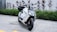 A sleek, modern electric scooter with a white and black color scheme, parked on a paved area next to green shrubs and red-tinted plants. The design is angular with a prominent front light and visible handlebars.