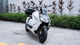 A sleek, modern electric scooter with a white and black color scheme, parked on a paved area next to green shrubs and red-tinted plants. The design is angular with a prominent front light and visible handlebars.