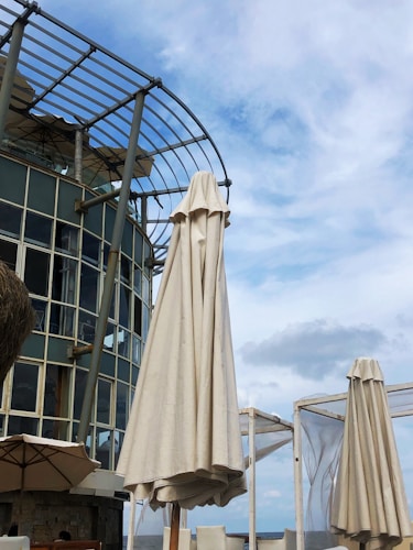 A tall, closed beige umbrella stands in front of a modern glass and metal building with a curved design. The sky is bright with scattered clouds, and there are other umbrellas and outdoor seating visible, suggesting a patio or open-air dining area.