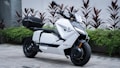 A sleek, modern scooter with a white body and black accents parked on a stone pavement. The scooter features sharp, angular design and is equipped with a storage compartment at the back. Several plants with red and green foliage are arranged behind the scooter against a white wall.