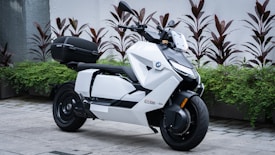A sleek, modern scooter with a white body and black accents parked on a stone pavement. The scooter features sharp, angular design and is equipped with a storage compartment at the back. Several plants with red and green foliage are arranged behind the scooter against a white wall.
