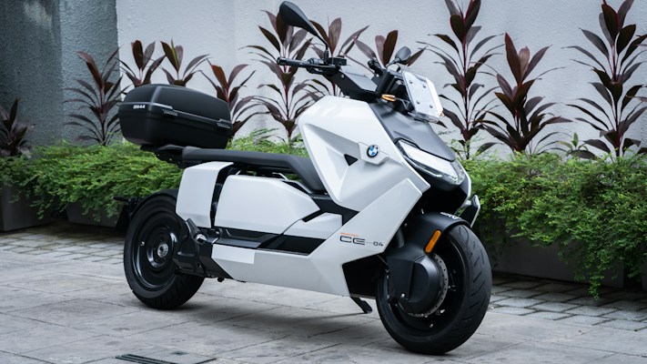 A sleek, modern scooter with a white body and black accents parked on a stone pavement. The scooter features sharp, angular design and is equipped with a storage compartment at the back. Several plants with red and green foliage are arranged behind the scooter against a white wall.