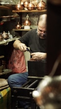 A craftsman is focused on his work in a workshop filled with copperware. He is seated, using a tool on a piece of metal with several copper pots and items surrounding him. Shelves and surfaces are cluttered with various tools and finished goods.