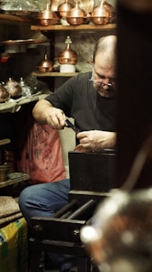 A craftsman is focused on his work in a workshop filled with copperware. He is seated, using a tool on a piece of metal with several copper pots and items surrounding him. Shelves and surfaces are cluttered with various tools and finished goods.