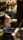 A craftsman is focused on his work in a workshop filled with copperware. He is seated, using a tool on a piece of metal with several copper pots and items surrounding him. Shelves and surfaces are cluttered with various tools and finished goods.