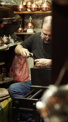 A craftsman is focused on his work in a workshop filled with copperware. He is seated, using a tool on a piece of metal with several copper pots and items surrounding him. Shelves and surfaces are cluttered with various tools and finished goods.