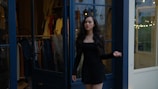 A woman with long dark hair is standing in front of a clothing store. She is wearing a black dress and appears to be looking confidently to the side. The store has glass doors with various clothing items displayed behind her.