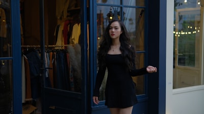 A woman with long dark hair is standing in front of a clothing store. She is wearing a black dress and appears to be looking confidently to the side. The store has glass doors with various clothing items displayed behind her.