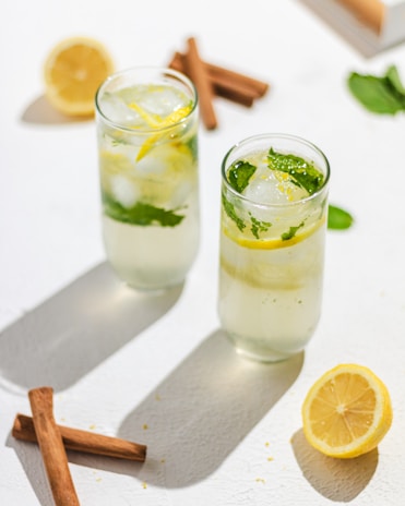 Two tall glasses filled with a refreshing beverage containing ice cubes, lemon slices, and fresh mint leaves. The glasses are placed on a white surface with natural light casting shadows. In the background, there are cinnamon sticks and a halved lemon, adding to the fresh and summery feel of the composition.