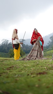 Two women dressed in traditional clothing are standing on a grassy field. One woman is wearing a red shawl and the other a yellow skirt with a beige shawl. They are engaged in an activity involving a large patterned cloth spread out on the ground. The sky is overcast, and the background features lush green hills and sparse trees.