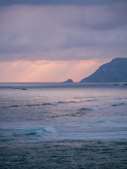 The image features a serene ocean view with gentle waves rolling towards the shore. A distant, rocky coastline is visible under a cloud-filled sky tinged with soft hues of pink and purple from the setting or rising sun.
