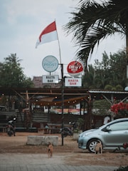 A small outdoor bar with a rustic and relaxed atmosphere, featuring a red and white flag on a pole. There are signs for Prost Beer and Draft Beer above the entrance, and two dogs are visible on the ground nearby. A silver car is parked to the right, with tropical plants and trees in the background. The area is decorated with string lights, adding a casual and inviting feel.