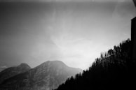 A black and white photograph captures a mountainous landscape with tall, rugged peaks and a dense forest covering the slopes. The sky appears partly cloudy, adding a dramatic contrast to the scene.