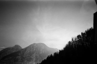 A black and white photograph captures a mountainous landscape with tall, rugged peaks and a dense forest covering the slopes. The sky appears partly cloudy, adding a dramatic contrast to the scene.