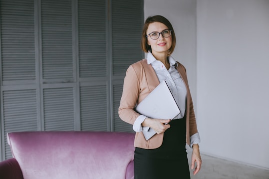A person stands confidently indoors, holding a closed laptop against their side. They are dressed in professional attire with a light blazer over a white shirt and dark skirt. The background includes a soft purple armchair and a backdrop of gray shutter doors.