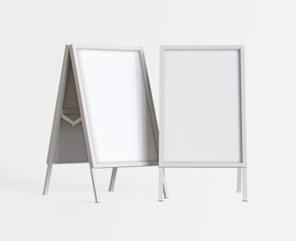 Two blank, white sandwich boards stand on a plain background. Each board has a sturdy, rectangular metal frame, supported by angled legs. The boards are positioned at slight angles to each other.