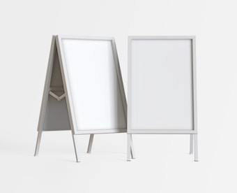 Two blank, white sandwich boards stand on a plain background. Each board has a sturdy, rectangular metal frame, supported by angled legs. The boards are positioned at slight angles to each other.
