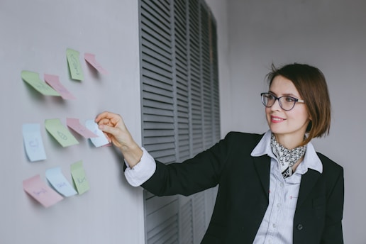 A woman wearing glasses and a black blazer is arranging colorful sticky notes on a wall. She appears focused and is smiling slightly. The background includes a modern, minimalistic design with a louvered panel.