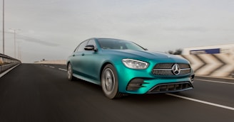 A sleek teal-colored Mercedes-Benz sedan is driving on a highway with a modern and aerodynamic design. The vehicle's front grille and headlights are prominently featured as it moves at a high speed. The road appears wide and smoothly paved, with visible lane markings. The background shows a blurred landscape, suggesting motion, under a clear sky with minimal clouds.