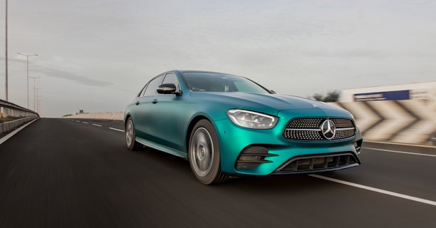 A sleek teal-colored Mercedes-Benz sedan is driving on a highway with a modern and aerodynamic design. The vehicle's front grille and headlights are prominently featured as it moves at a high speed. The road appears wide and smoothly paved, with visible lane markings. The background shows a blurred landscape, suggesting motion, under a clear sky with minimal clouds.
