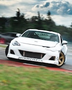 A white sports car is drifting on a racetrack, with noticeable decals and modifications. The car is angled to one side, suggesting high-speed movement. It is set against a blurred background of trees and sky, indicating motion and speed.