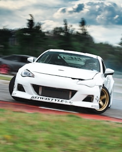 A white sports car is drifting on a racetrack, with noticeable decals and modifications. The car is angled to one side, suggesting high-speed movement. It is set against a blurred background of trees and sky, indicating motion and speed.