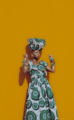 A person wearing a vibrant dress with bold, circular patterns stands confidently against a mustard yellow background. The dress features green and black concentric designs, and the person accessorizes with matching bracelets and a head wrap.