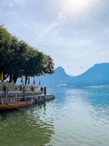 A serene lakeside with clear blue water reflecting the sky. A wooden boat is moored alongside a stone dock lined with tables and chairs shaded by large trees. Distant green hills and mountains create a picturesque backdrop.