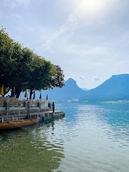 A serene lakeside with clear blue water reflecting the sky. A wooden boat is moored alongside a stone dock lined with tables and chairs shaded by large trees. Distant green hills and mountains create a picturesque backdrop.