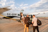 Several people are boarding a small aircraft on a tarmac under a partly cloudy sky. The plane has the registration 5X-BXW and belongs to a company. Passengers are carrying bags as they ascend the stairs.