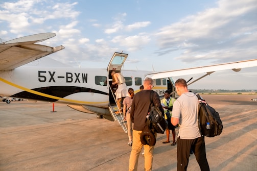 Several people are boarding a small aircraft on a tarmac under a partly cloudy sky. The plane has the registration 5X-BXW and belongs to a company. Passengers are carrying bags as they ascend the stairs.