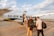 Several people are boarding a small aircraft on a tarmac under a partly cloudy sky. The plane has the registration 5X-BXW and belongs to a company. Passengers are carrying bags as they ascend the stairs.