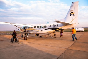 A small aircraft with the registration 5X-BXW and AeroLink branding is stationed on a tarmac. Several people are seen around the plane, some carrying luggage and others walking towards it. The surrounding area appears to be an airport with a wide view of the sky and distant landscape.