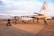 A small aircraft with the registration 5X-BXW and AeroLink branding is stationed on a tarmac. Several people are seen around the plane, some carrying luggage and others walking towards it. The surrounding area appears to be an airport with a wide view of the sky and distant landscape.
