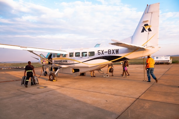 A small aircraft with the registration 5X-BXW and AeroLink branding is stationed on a tarmac. Several people are seen around the plane, some carrying luggage and others walking towards it. The surrounding area appears to be an airport with a wide view of the sky and distant landscape.