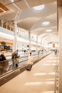 A spacious modern airport terminal with a high, curved ceiling featuring circular lights. Several travelers walk along a moving walkway, some pulling suitcases. The area is brightly lit with natural light from large windows, creating a clean and airy atmosphere. Shops and gates are visible in the background.