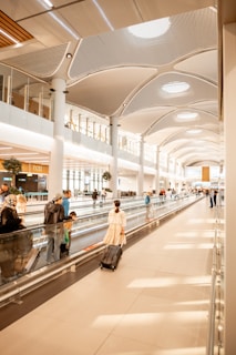 A modern airport terminal with large glass windows and travelers walking with rolling suitcases.